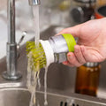 Person holding a multifunctional kitchen scrub brush with soap dispenser under running water in a sink.