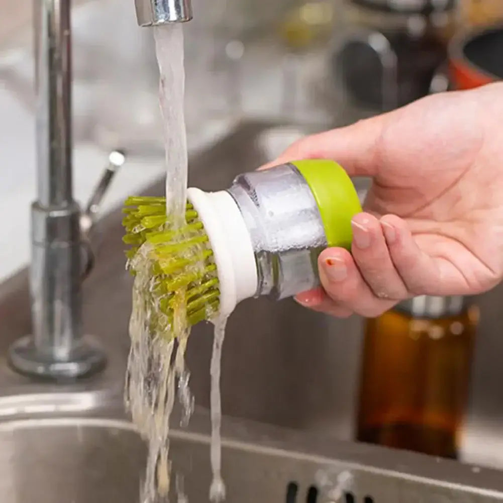 Person holding a multifunctional kitchen scrub brush with soap dispenser under running water in a sink.